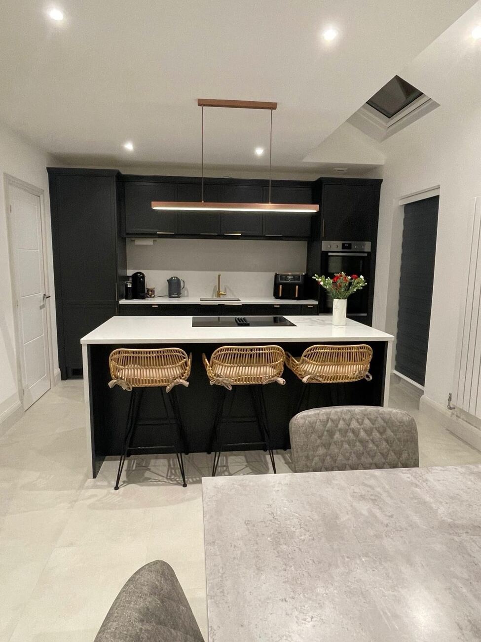 Black kitchen with a white-topped island, three rattan bar stools and a copper pendant light